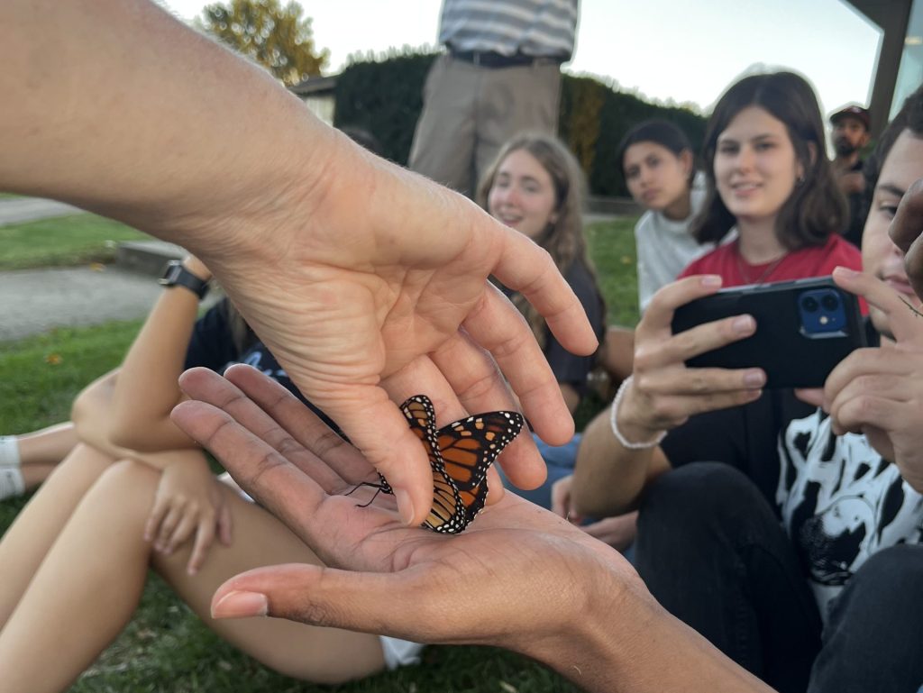 A monarch butterfly is gently placed into an open hand, ready for release, as a group of young adults watch